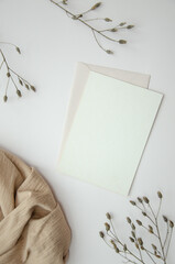 Blank paper card on white background with dried plants and wavy beige fabric, top view. Boho mock-up scene with greeting card template