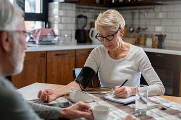 An elderly woman checks her blood pressure at home using a digital monitor while an elderly man...