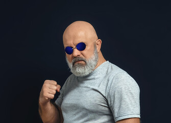 Menacing bald man with gray beard in dark glasses looks at camera and raises his fist. Cruelty.