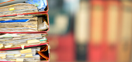 stack of messy file folders, blurred bookshelf in the back,red tape, bureaucracy,beat the clock...