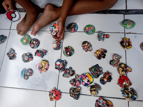 A child's hands carefully arrange colorful, round tazos on a tiled floor, with a variety of tazos scattered around them. The tazos feature various cartoon characters and patterns, and a single Pokemon