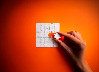A hand carefully places the final white jigsaw piece into a square puzzle against a vibrant orange backdrop, symbolizing completion and problem-solving.