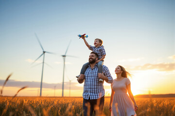 Family enjoying sunset in a wind farm field with child holding toy airplane.