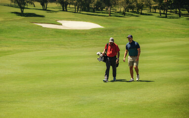 Two male golfers walking together on a sunny fairway, carrying golf bags near a sand bunker