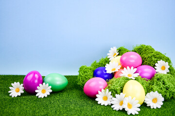 Easter composition with brightly colored eggs lying on fresh green moss decorated with white daisies, against a background of green grass and blue sky.