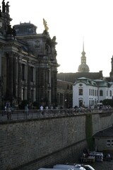 Fototapeta premium Historic architecture with church spire at sunset, people walking along riverside promenade, symbolizing culture, heritage, and timeless urban charm.