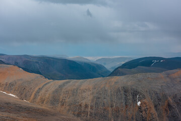 Dramatic layered aerial view above large sharp ridge to green alpine deep valley among big cliffs far away in rain under gray cloudy sky. High rocky mountain silhouettes on horizon in rainy low clouds