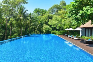 Fototapeta premium Infinity Pool Tropical Getaway - Stunning infinity pool overlooking lush tropical landscape, perfect for relaxation and luxury vacation. Sun loungers and umbrellas provide shade and comfort.