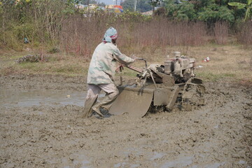 The farmer is plowing the muddy field with the power tiller and he has completely covered by the mud