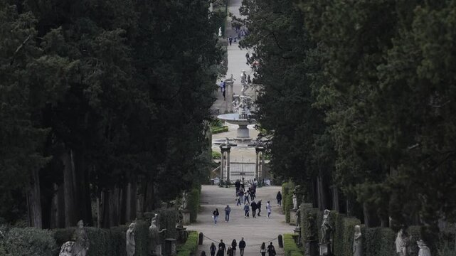 A group of people walk down a path in a park. The path is lined with trees and statues
