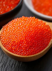 Close up view of vibrant red fish eggs in a wooden spoon, set against a dark background. The eggs are glistening and appear fresh