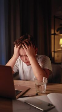 Stressed young boy holding head in frustration sitting at desk in cozy evening room emotionally reacting to laptop screen overwhelmed by online task game or remote schoolwork