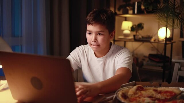 Cute little multiethnic boy at night working on homework typing browsing having distance education on laptop computer and eating pizza fast food at home