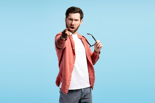 Aggressive bearded young man holding eyeglasses pointing at camera on blue background
