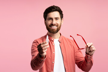 Smiling bearded man holding eyeglasses holding ophthalmology eyedropper, isolated on pink