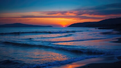 Obraz premium Serene coastal sunset with deep blue and orange hues over calm ocean waves and silhouetted hills creating a peaceful evening atmosphere.