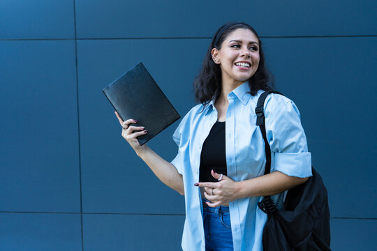 Young woman smiling while holding a black notebook and wearing a light blue shirt, black top, jeans, and a backpack standing against a modern dark blue wall outdoors. 