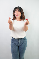 Beautiful young asian woman with wavy hair wearing white shirt is showing love heart hand gesture, isolated over white background.