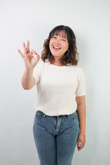 Beautiful young asian woman with wavy hair wearing white shirt is showing okay hand gesture, isolated over white background.