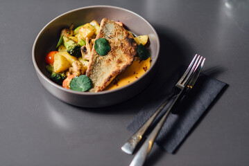 Crispy seabass fillet with roasted vegetables, creamy bisque sauce, and fresh herbs, elegantly plated in a dark ceramic bowl on a stylish minimalistic table setting