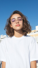 Portrait of a confident young woman with curly hair wearing sunglasses and a white t-shirt, posing against a clear blue sky