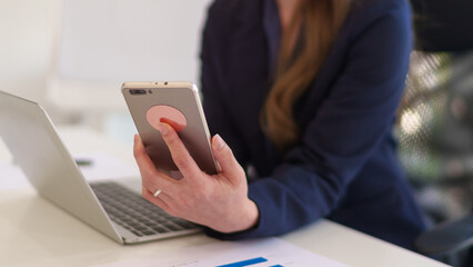 Young Asian businesswoman holding a smartphone while working on a laptop in a modern office with a presentation board