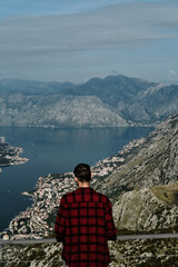 Fototapeta premium A young Caucasian man in a red plaid shirt stands on an observation deck overlooking the Bay of Kotor. Rear view. The southernmost fjord in the Adriatic Sea, Montenegro. Travel concept