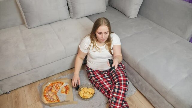 A woman in pajamas sits on the floor near the couch, holding a slice of pizza and a TV remote. On the rug in front of her are a pizza box and a bowl of chips