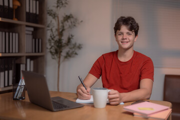 Happy teenager studying at home, using a laptop while taking notes and sipping coffee, smiling and enjoying the learning experience in a cozy, inviting room