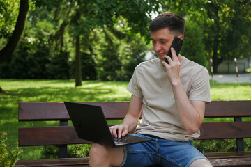 Fototapeta premium The man, working remotely in the park, uses his laptop while chatting on the phone. He blends freelancing with the convenience of online learning, surrounded by the peaceful outdoor environment.