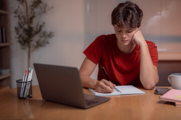 Young student taking notes in a notebook while studying at home, focused on a laptop at his desk....