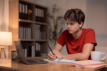 Young man writing in a notebook while focusing on a laptop, participating in an online class or e-learning course, studying at home during the evening hours