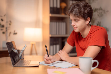 Fototapeta premium Young man taking notes in a notebook while using a laptop computer at home, studying or working late at night in a cozy setting with a lamp and a mug on the table