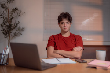 Young man in a red shirt sitting at a desk with a laptop, notebook, and smartphone. A coffee mug is nearby, suggesting a focused and productive work environment