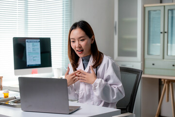 Asian female doctor wearing lab coat speaking with patient via video conference on laptop in clinic...