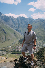 Smiling man in the mountains of Georgia with Kazbegi village below, showing a hand gesture against a backdrop of green valleys and rugged peaks under a sunny sky
