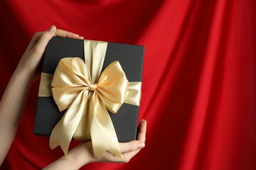 Woman holding gift box with beautiful bow against red curtain, closeup. Space for text