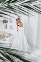 Fototapeta premium Cute girl standing with her arms crossed in a white dress with a train. Photo through palm tree leaves