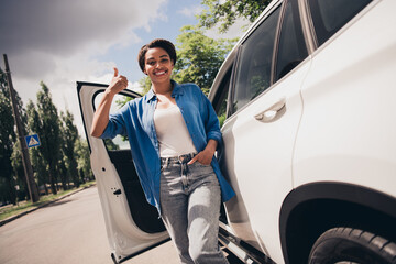 Confident young female driver gives thumbs up near car on sunny day, representing urban travel and freedom