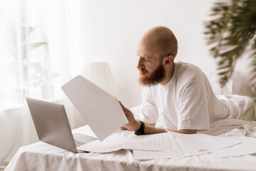 Businessman on bed, using laptop and paper documents. man working in bedroom. Businessperson stayed at home.