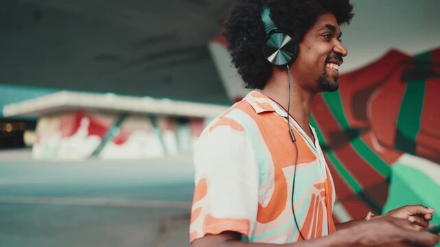 Close-up of cheerful young African American man wearing shirt listening to music in headphones and dancing on urban city background. Lifestyle concept. Slow motion