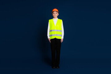 Mature businesswoman in safety gear with helmet and vest standing confidently against dark blue background.