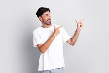 Cheerful Young Man in Casual White T-Shirt Pointing with Both Hands Indoors on a Light Grey Background