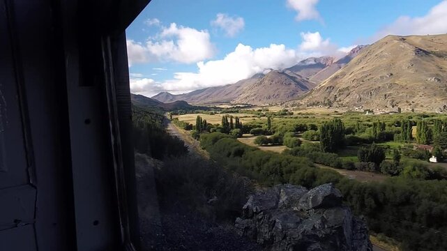 From the window of La Trochita train, Esquel Valley and the Andes unfold in an impressive landscape with clouds. The camera vibrates slightly due to the movement of the old tracks.