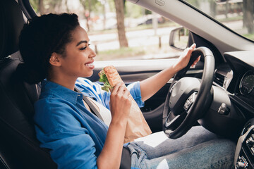 Cheerful young woman driving car and enjoying a sandwich during a sunny day in the city