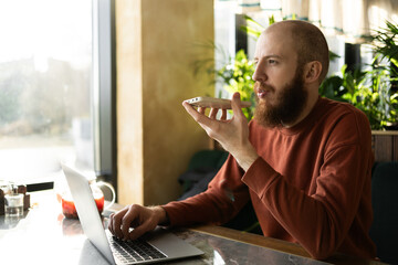 Young businessman with computer in modern green cafe, recording audio message for client on smartphone. voicemail