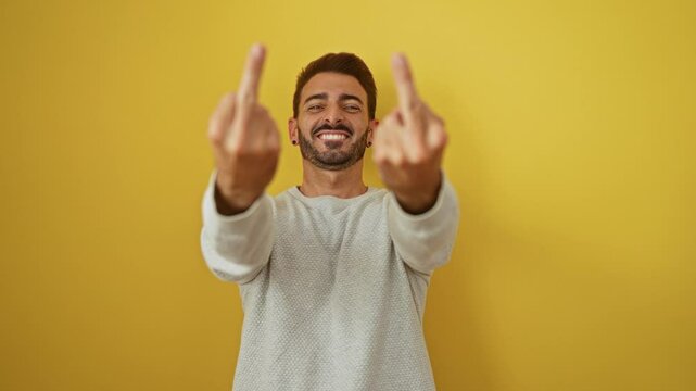 Hispanic man smiling against yellow background showing offensive gesture with both hands in casual white sweater expressing humor and rebellious attitude in a portrait setting