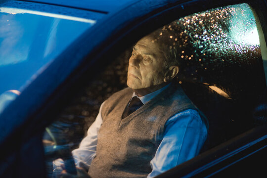 Senior man sitting in car, lost in thought during a rainy evening