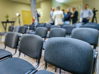 Rows of empty black chairs in a conference room with a blurred background of people engaged in...