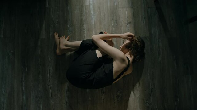 High angle of miserable young woman in black chemise experiencing panic attack lying on floor hugging her knees